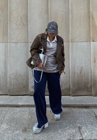 Καφέ δερμάτινο μπουφάν πάνω από ένα ριγέ πόλο, ναυτικές φόρμες και ασημένια πλατφόρμα γαλότσες. Κρατάει μια μικρή μεταλλική τσάντα. Ουδέτερο φόντο.