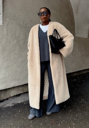 Mujer con abrigo teddy beige de gran tamaño, pantalones grises de pierna ancha, suéter gris superpuesto sobre camisa blanca, sosteniendo un bolso negro bajo el brazo.
