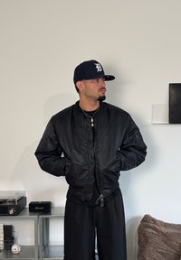 Chaqueta bomber negra con textura brillante, cierre de cremallera y bolsillos laterales, usada sobre una camiseta negra. La persona lleva una gorra oscura.