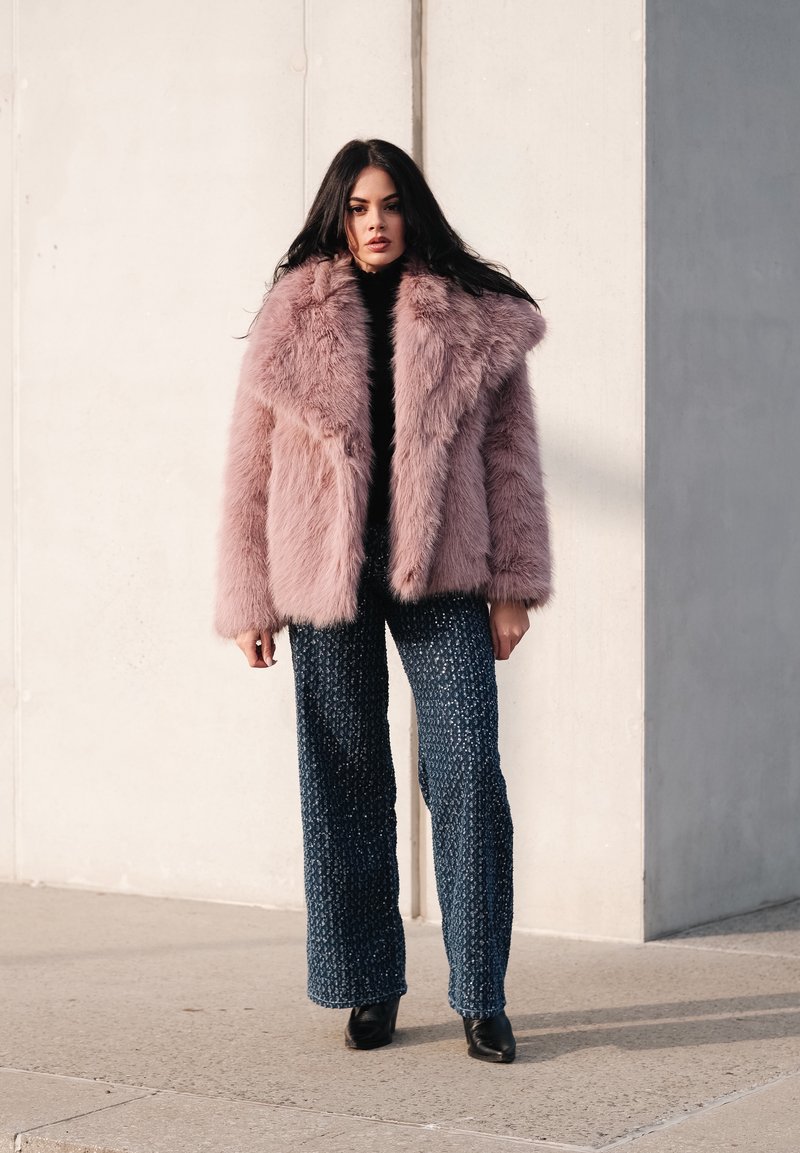 Manteau en fausse fourrure rose clair, à larges revers crantés, porté sur un col roulé noir. Pantalon large à motifs bleus et bottes noires complètent le look.