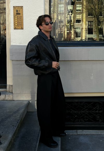 Chaqueta bomber de cuero negra con puños acanalados, camisa negra ajustada, pantalones negros sueltos y zapatos negros. Las gafas de sol completan el look.