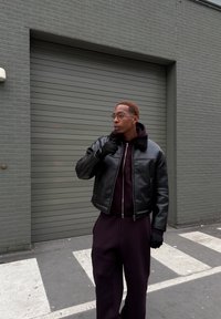 Veste bomber en cuir noir avec un col en fourrure, portée sur un sweat à capuche foncé. Le modèle porte des gants noirs, debout contre un mur gris.