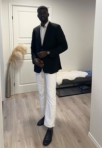 Schwarze Sakko über einem weißen Hemd, kombiniert mit weißen Hosen und schwarzen Schuhen. Merkmale einer runden Uhr am linken Handgelenk. Schlichter Hintergrund.