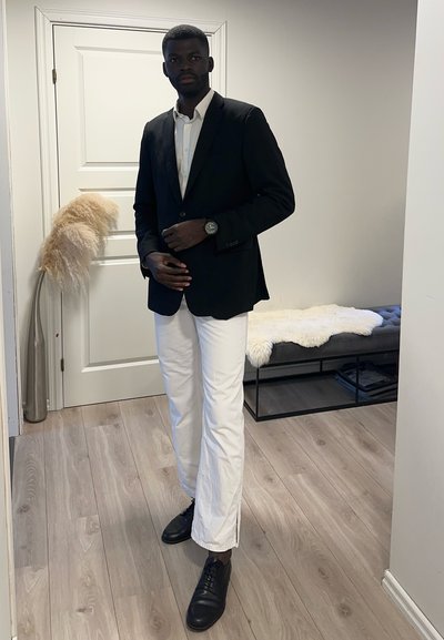 Chaqueta de traje negra sobre una camisa blanca, combinada con pantalones blancos y zapatos negros. Lleva un reloj redondo en la muñeca izquierda. Fondo sencillo.