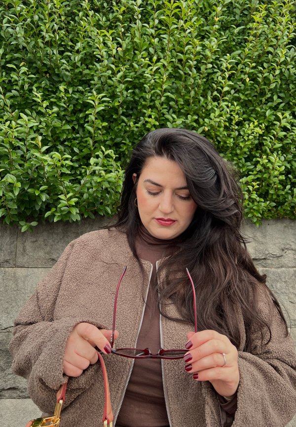 Braune Fleece-Jacke mit kontrastierendem silbernen Reißverschluss, lange dunkle Haare, hält rote Sonnenbrille, steht vor einer grünen Hecke und einer Steinmauer.