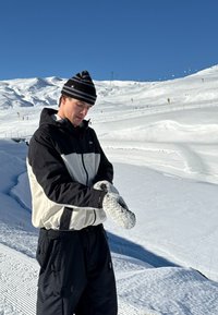 Černobílá zateplená bunda s kapucí, černé kalhoty a šedé pletené rukavice. Sněhová krajina a modrá obloha v pozadí.