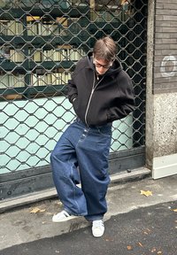 Ung person i svart hettegenser, store blå jeans og hvite joggesko står utendørs ved en lukket metallport med hendene i lommene.