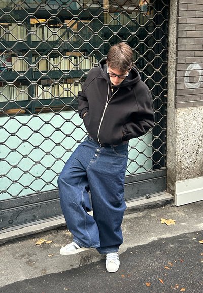 Persona joven con sudadera negra con capucha, vaqueros azules holgados y zapatillas blancas, de pie al aire libre junto a una puerta metálica cerrada con las manos en los bolsillos.
