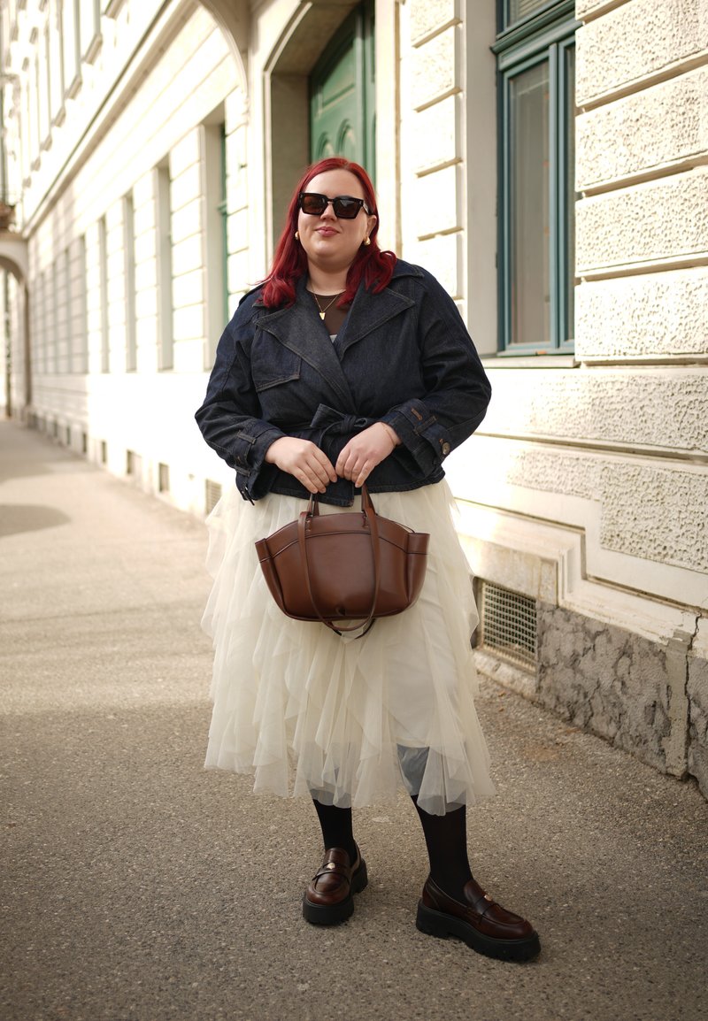 Žena s červenými vlasmi, ktorá nosí slnečné okuliare, tmavú bundu, bielu vrstvenú sukňu a hnedé mokasíny, drží hnedú kabelku na mestskom chodníku.