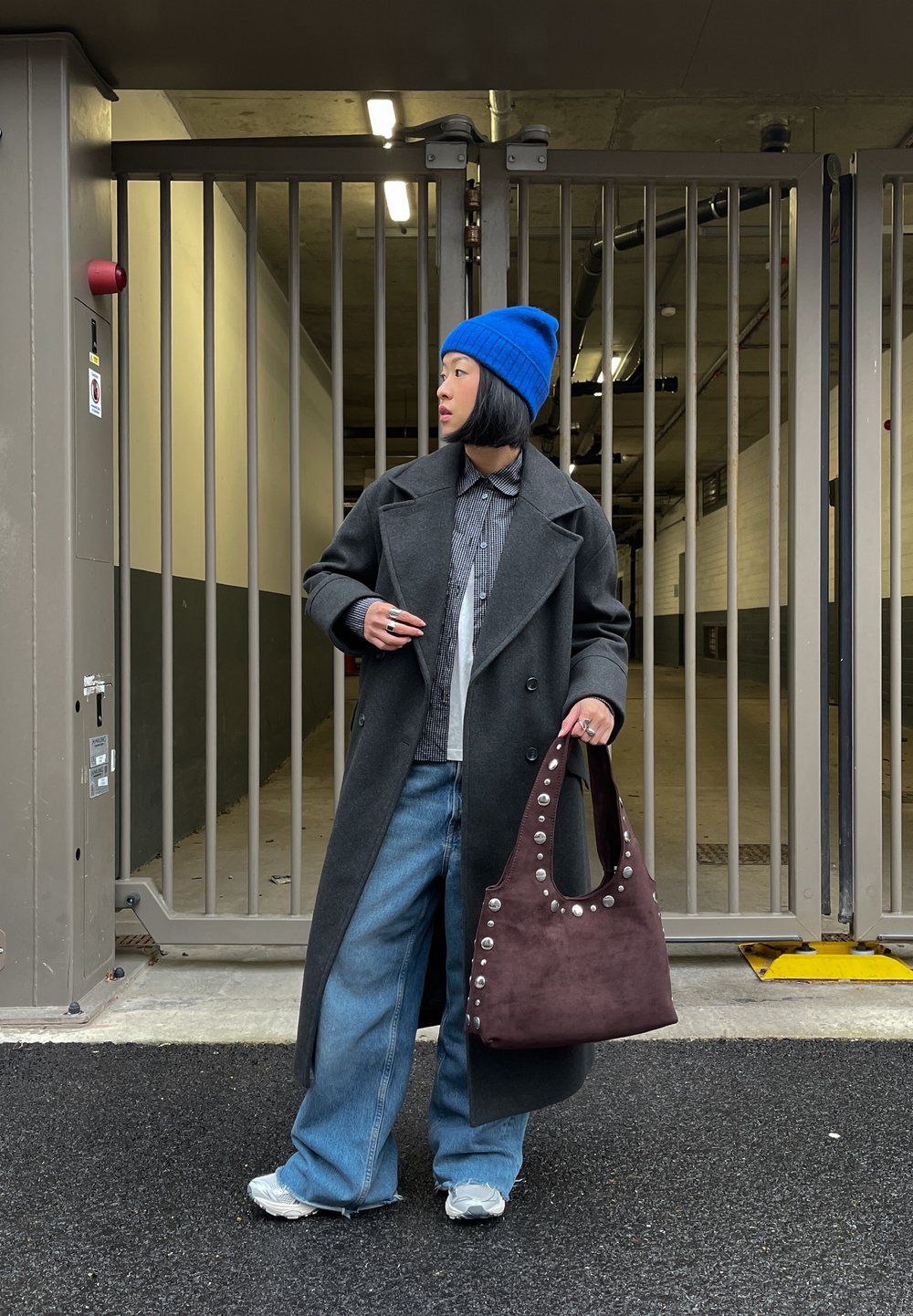 Personne portant un bonnet bleu, un manteau gris foncé, une chemise à carreaux, un jean ample et des baskets blanches, tenant un sac à main marron clouté près d'une grille métallique.