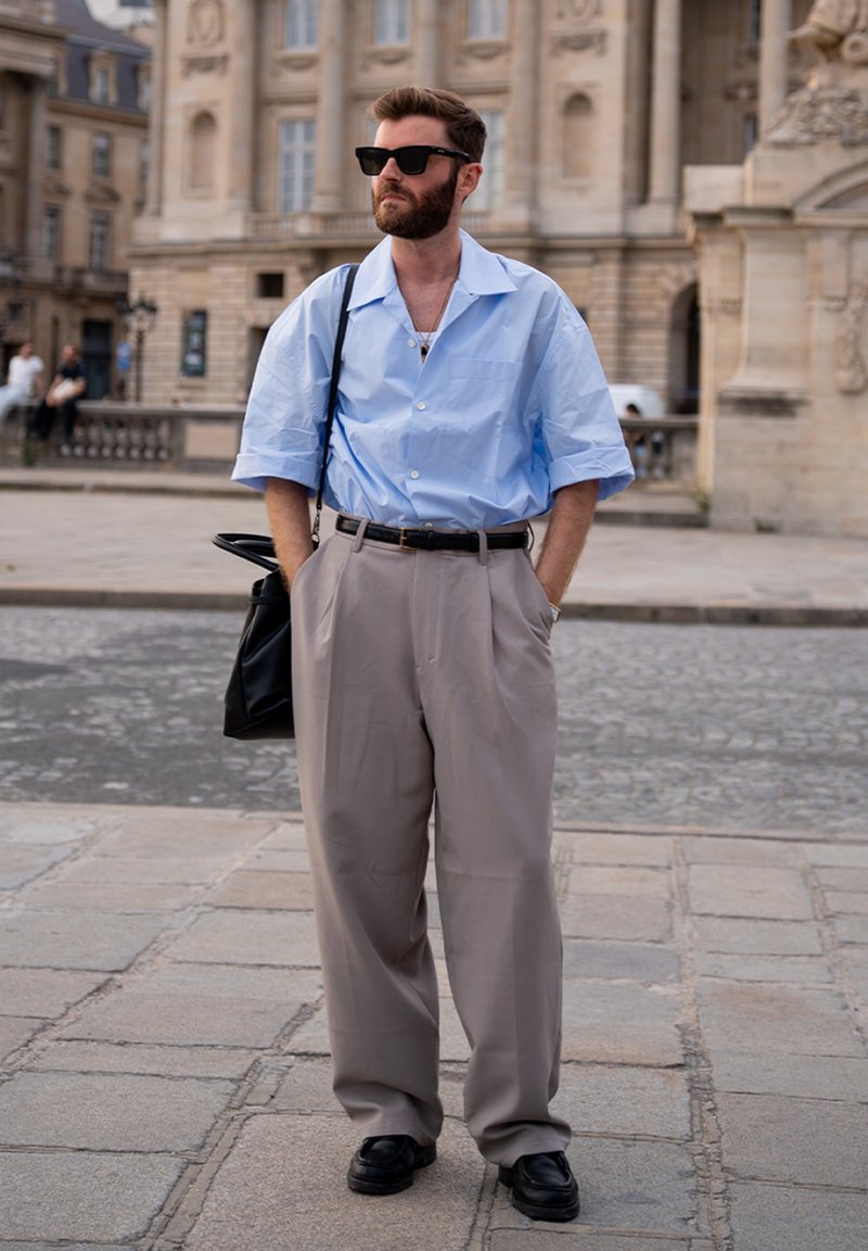 Homme barbu portant des lunettes de soleil, une chemise à manches courtes bleu clair, un pantalon large beige, des chaussures noires, et portant un sac à épaule noir dans un contexte urbain.