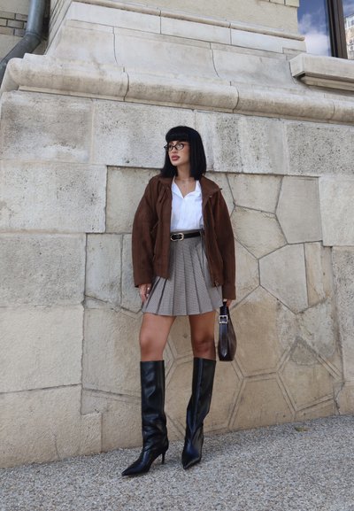 Chaqueta de ante marrón, camisa blanca, falda de cuadros gris con pliegues, botas de cuero negras hasta la rodilla y un pequeño bolso marrón, contra una pared de piedra.