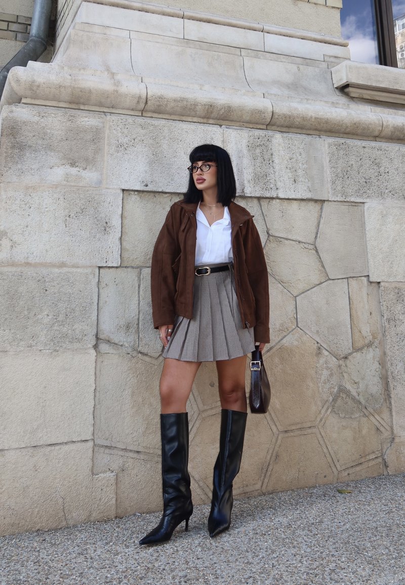 Veste en daim marron, chemise blanche, jupe plissée grise à carreaux, bottes en cuir noires jusqu'aux genoux et petit sac à main marron, devant un mur en pierre.