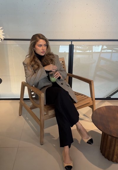 Mujer con blazer de cuadros y pantalones negros sentada en una silla de madera bebiendo una bebida verde con pajita en un espacio interior moderno.