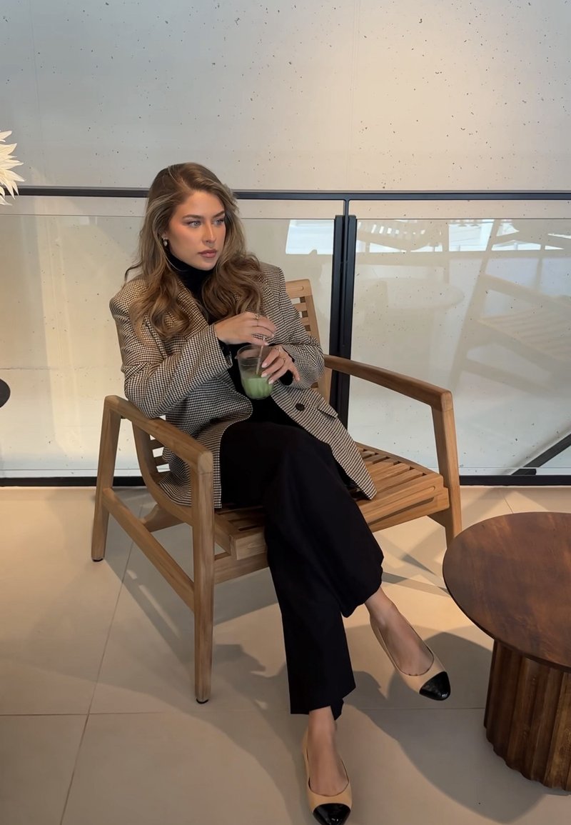 Kvinna i rutig kavaj och svarta byxor sitter på en trästol och sörplar en grön dryck med sugrör i ett modernt inomhusutrymme.