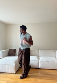 Mann mit ärmellosem Pullover und weiten braunen Hosen steht auf einem Holzboden neben einem weißen Ecksofa mit Leopardenmuster-Kissen.