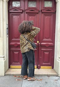 Oversized shirt met luipaardprint, zwarte rechte jeans met rafelranden en flats met luipaardprint. De achtergrond toont een bordeauxrode dubbele deur.