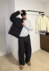 Veste matelassée noire tenue partiellement ouverte sur un t-shirt à manches longues gris, un pantalon large noir, et des chaussures marron. Cadre intérieur minimaliste.