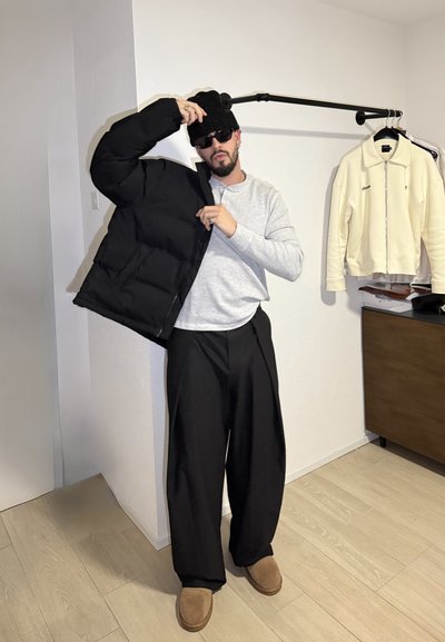 Chaqueta negra acolchada parcialmente abierta sobre una camiseta de manga larga gris, pantalones anchos negros y calzado marrón. Entorno interior minimalista.