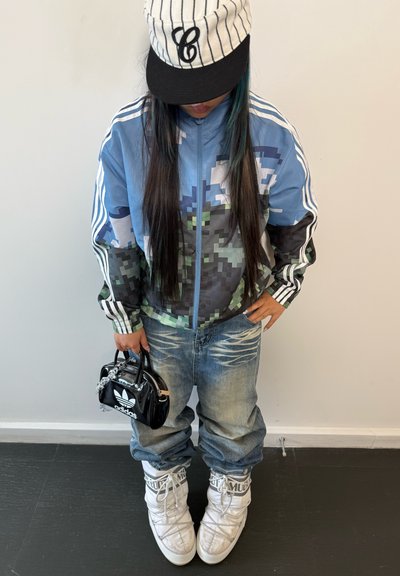 Chaqueta de camuflaje azul y negra con cremallera, jeans de mezclilla azul claro, zapatillas altas blancas y un pequeño bolso negro con una cadena plateada.