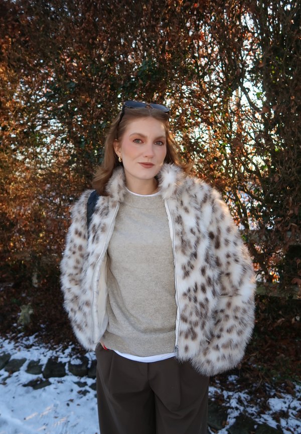 Vrouw die een bontjas met vlekken draagt, een beige trui en donkere broek, staand in de buitenlucht bij bladloze struiken met plekken sneeuw op de grond.