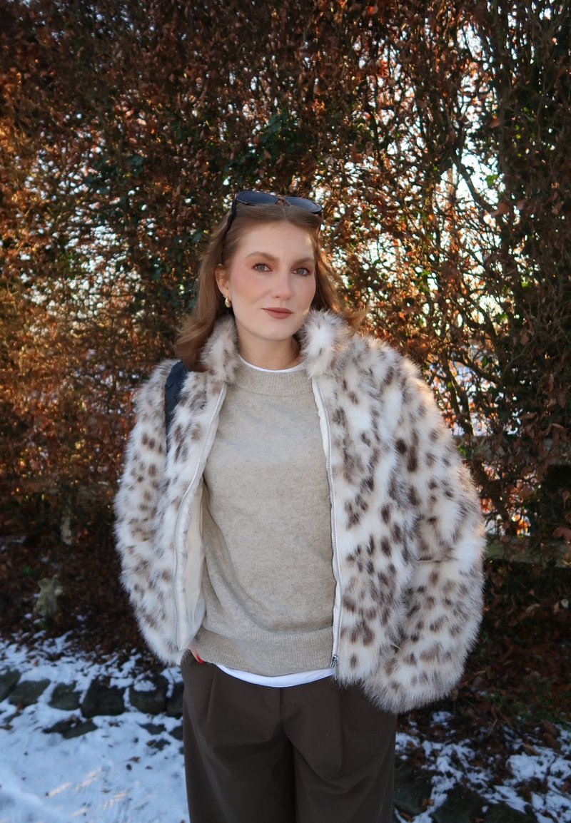 Mujer con abrigo de piel con manchas, suéter beige y pantalones oscuros, de pie al aire libre cerca de arbustos sin hojas con parches de nieve en el suelo.