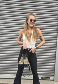Črne visoke flare jeans hlače, bela zavita bluza in majhna torbica z vzorcem. Sončna očala in vidne tetovaže na rokah.
