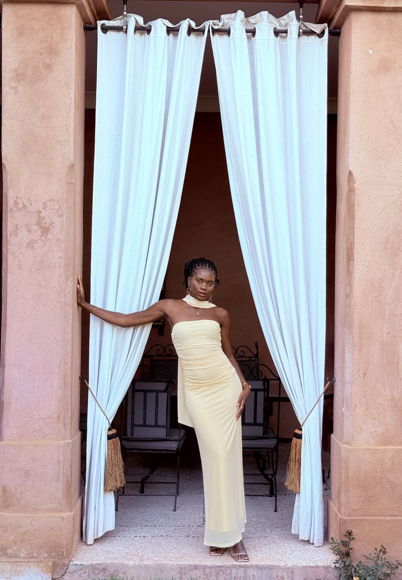 Robe jaune sans bretelles avec un drapé sur le côté, fabriquée en tissu lisse. Des rideaux blancs encadrent la scène, avec des chaises noires visibles derrière.