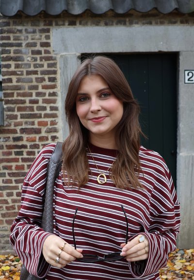 Camiseta de rayas largas en granate y blanco, sujetando gafas de sol negras, con joyas doradas, frente a una pared de ladrillo con una puerta verde.