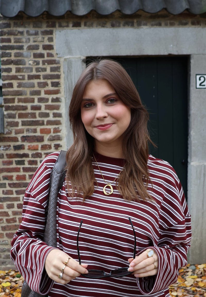 Chemise à manches longues rayée bordeaux et blanche, tenant des lunettes de soleil noires, portant des bijoux en or, posée devant un mur en briques avec une porte verte.