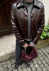 Bruine leren cropped jas met een glanzende afwerking, grijze kabeltrui, donkergrijze broek en een geweven bordeauxhandtas.