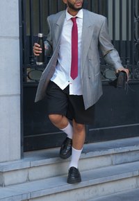 Γκρίζα υπερμεγέθη σακάκι, λευκό πουκάμισο με ανοιχτό γιακά, κόκκινη γραβάτα, μαύρες βερμούδες, μαύρα παπούτσια και λευκές κάλτσες. Κρατάει ένα μπουκάλι νερού και πορτοφόλι.