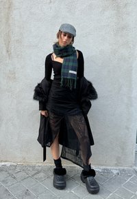 Μαύρη διαφανής φούστα με δαντελωτά σχέδια, μακρύ μαύρο παλτό, ριμπ πουκάμισο σε μαύρο χρώμα, καρό πράσινο κασκόλ, γκρι καπέλο και μαύρες φλάντζες πατούσες.