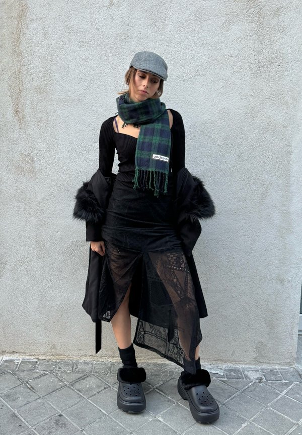 Jupe noire transparente avec des motifs en dentelle, long manteau noir, haut noir côtelé, écharpe à carreaux verte, casquette grise et chaussures noires en peluche à enfiler.