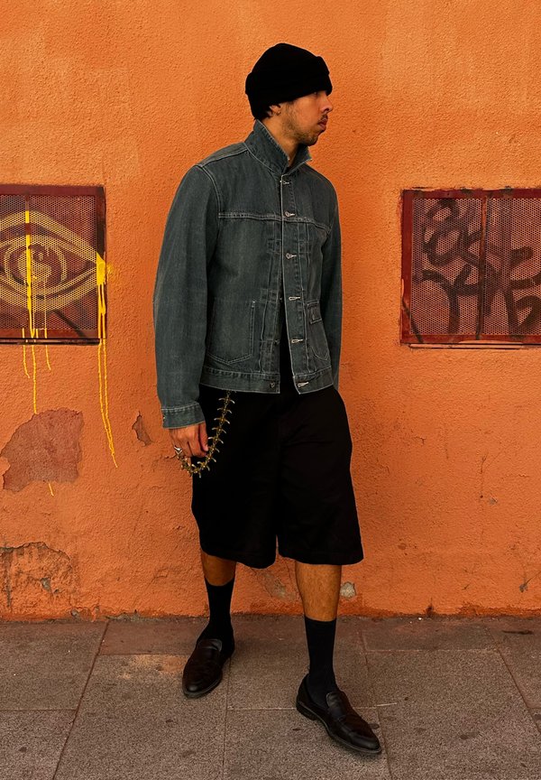 Denim jacket in blue-grey, black shorts, black socks, and black shoes. Accessories include a chain. The background features an orange wall and graffiti.