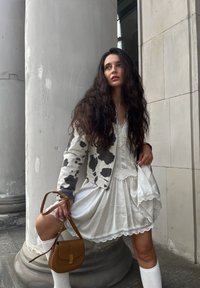 White dress with lace trim paired with a grey and black cow-print jacket. Brown handbag with a strap, knee-high white socks, textured hair.