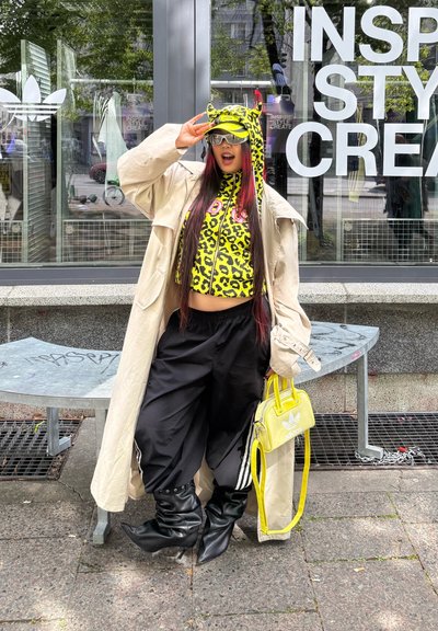 Modelo vistiendo un top corto de leopardo amarillo, pantalones holgados negros y un abrigo beige, sosteniendo un bolso amarillo, de pie en un entorno urbano.