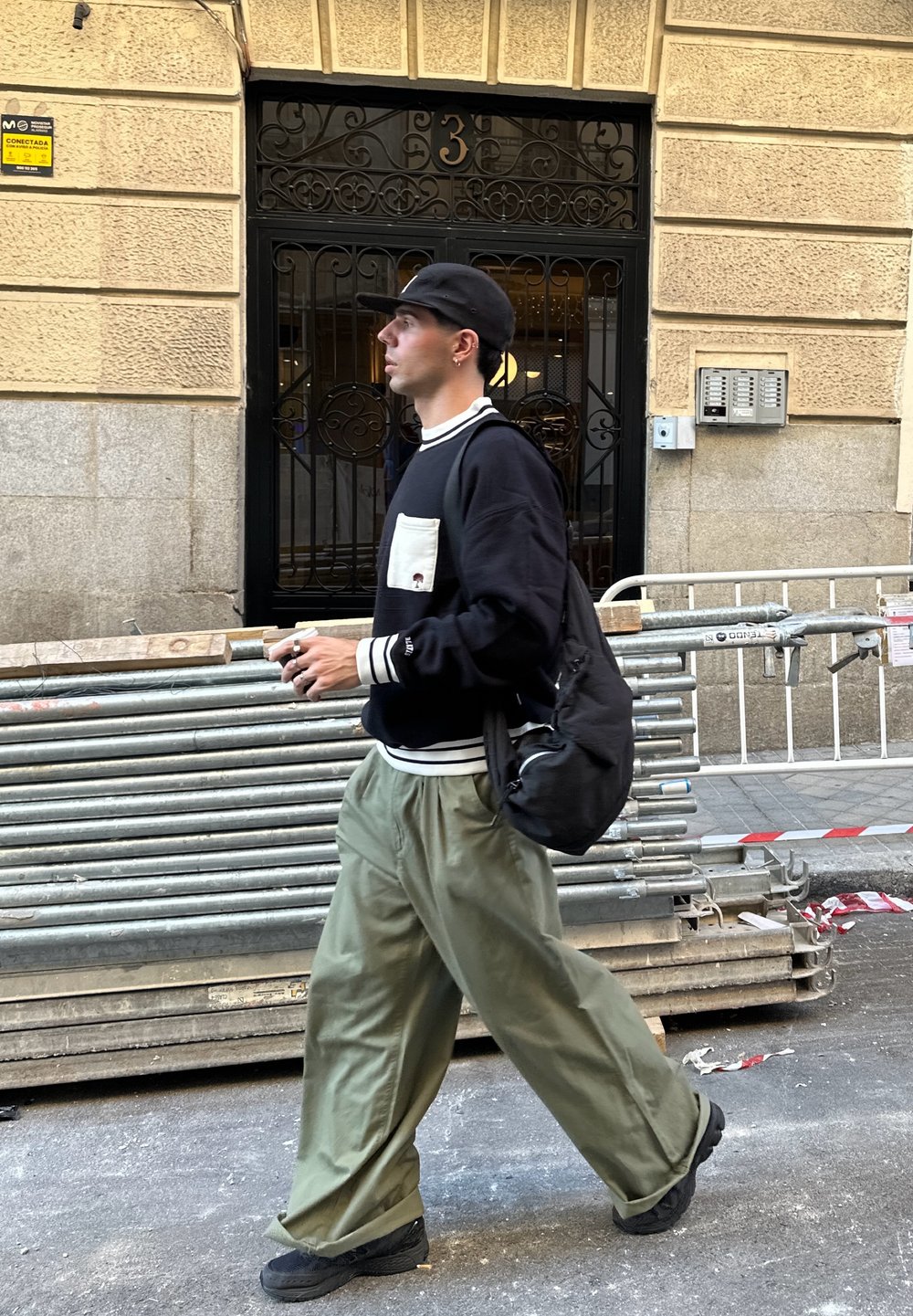 Hombre caminando junto a tuberías metálicas apiladas y un edificio con paredes de piedra amarilla, vistiendo una gorra negra, un suéter negro y pantalones verdes holgados.