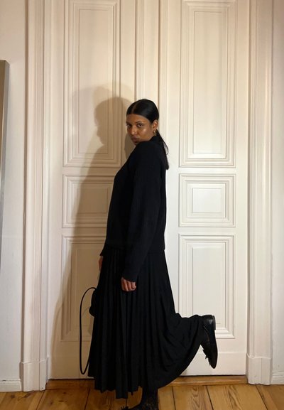 Suéter negro de canalé con mangas largas, combinado con una falda negra de pliegues fluida. Botines negros completan el atuendo. El fondo presenta paneles blancos.