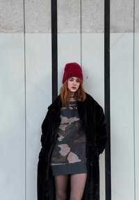 Čierny kabát z umelého kožušinového materiálu cez sivé šaty s maskáčovým vzorom, doplnené červenou pletenou čiapkou, na pozadí svetlosivej steny s vertikálnymi pruhmi.