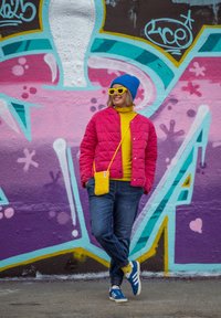 Ροζ καπιτονέ μπουφάν, κίτρινο πουλόβερ, μπλε σκουφάκι και γυαλιά ηλίου. Μπλε τζιν και παπούτσια αθλήσεως, κρατώντας μια κίτρινη τσάντα χιαστί. Φόντο με γκράφιτι.