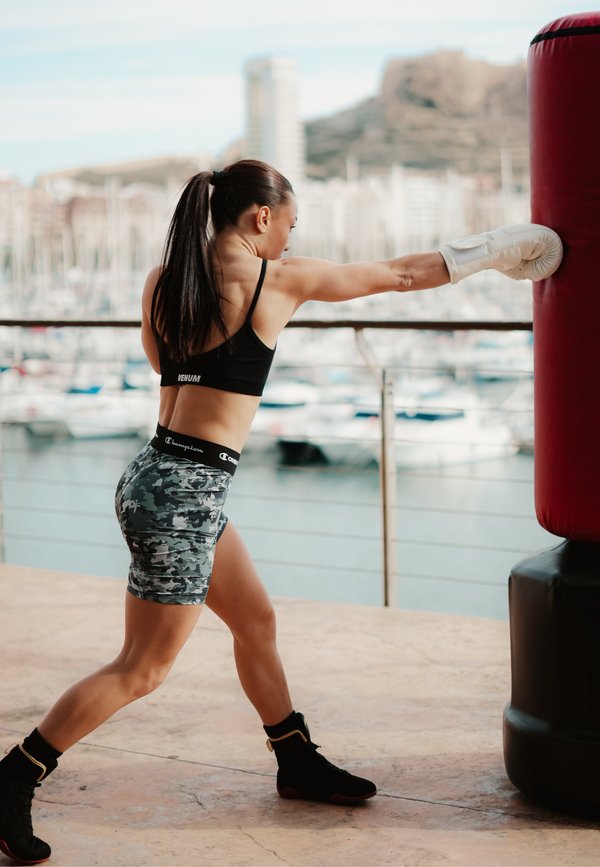 Femme en tenue de sport et gants de boxe donnant un coup de poing à un sac de frappe rouge sur une terrasse extérieure avec vue sur des bateaux et des bâtiments.