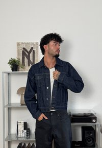 Jachetă de denim albastru închis cu un design scurt, având un guler ascuțit, închidere cu nasturi și două buzunare pe piept. Pur tată pe un cămașă deschisă.