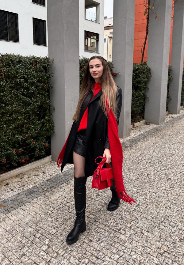 Cappotto doppiopetto nero, maglione rosso, pantaloncini di pelle nera, sciarpa rossa lavorata a maglia, stivali neri al ginocchio e piccola borsa rossa. Sentiero di ciottoli.