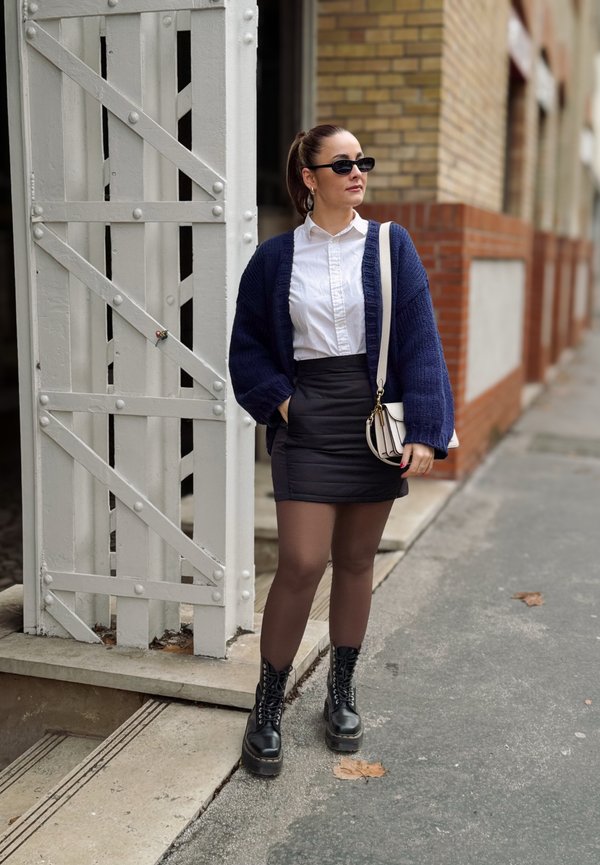 Navy gestrickte Strickjacke, weißes Hemd, schwarzer Minirock, braune Strumpfhosen und schwarze Schnürstiefel. Kleine beigefarbene Handtasche mit Akzentbeschlägen.