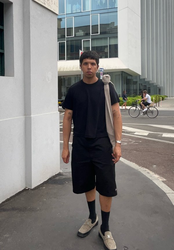 Black short-sleeve shirt and black shorts. Grey slip-on shoes. Light grey fabric draped over the shoulder. Urban setting, concrete background.