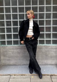 Schwarze, kurze Jacke über einem weißen Rollkragenpullover, kombiniert mit schwarzen Jeans mit weitem Bein und schwarzen Sneakern. Die Haare sind mit einem roten Haaraccessoire gestylt.