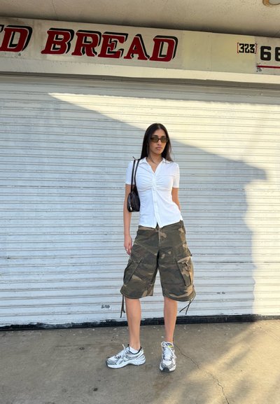 Camisa blanca de manga corta con fruncido en el frente, pantalones cortos cargo de camuflaje y zapatillas grises. Un bolso negro de hombro y gafas de sol polarizadas completan el look.