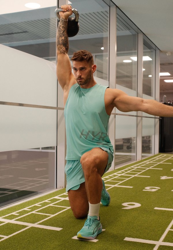 Un hombre vestido con ropa deportiva de color turquesa sostiene una pesa rusa por encima de la cabeza mientras hace una zancada en una pista cubierta de césped artificial.