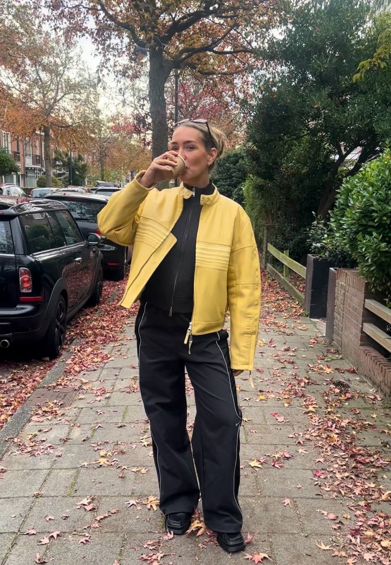 Vrouw in geel jack en zwarte broek drinkt uit een beker op een met bladeren bedekt stedelijk trottoir met geparkeerde auto's en bomen.